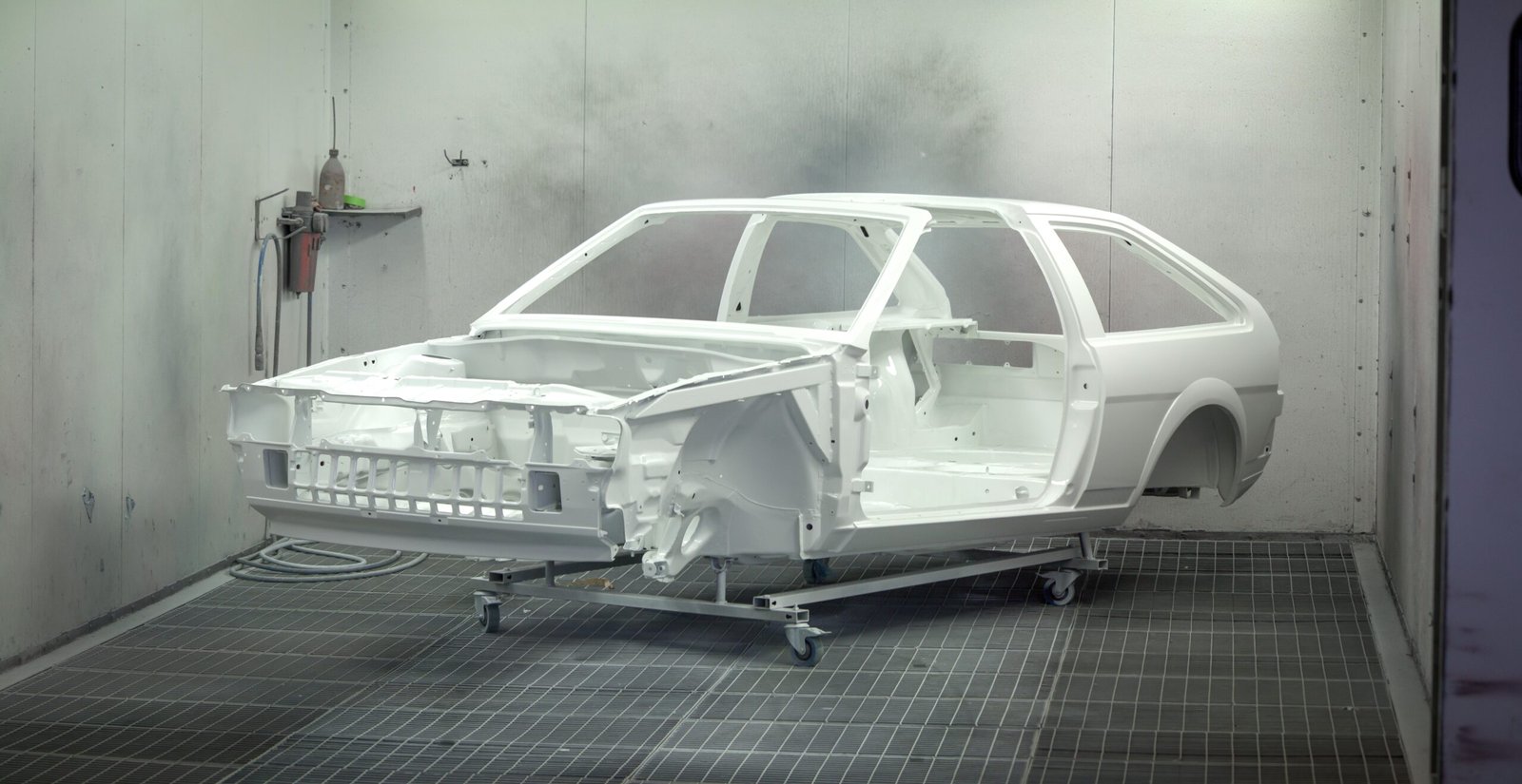 A vintage car frame being painted white in an industrial automotive paint booth.